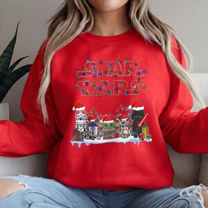 Pode incluir: Camisola vermelha com o texto "STAR WARS" em uma fonte festiva e iluminada. Abaixo, v&aacute;rios personagens de Star Wars s&atilde;o retratados usando gorros de Papai Noel e cercados por decora&ccedil;&otilde;es de Natal. Um design divertido e sazonal.