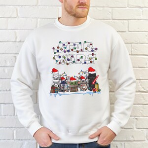 Pode incluir: Camisola branca com um design de Star Wars. O design apresenta personagens com gorros de Papai Noel e luzes de Natal que soletram "STAR WARS". Os personagens incluem Stormtrooper, R2-D2, Grogu e Darth Vader.