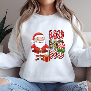 Op de afbeelding: Witte sweatshirt met een cartoon kerstman die knipoogt, een rood pak en hoed draagt en een cadeau vasthoudt. De woorden "HO HO HO" zijn gedrukt in rode stippen, luipaardprint en rood-witte strepen, met een groene strik.