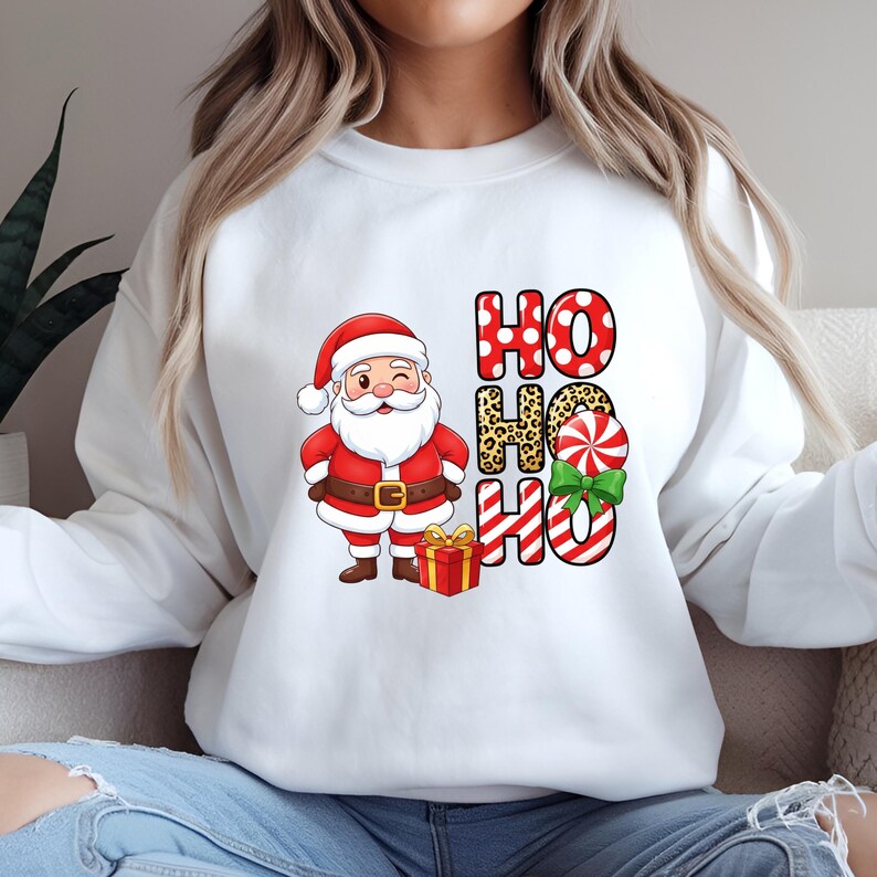 Op de afbeelding: Witte sweatshirt met een cartoon kerstman die knipoogt, een rood pak en hoed draagt en een cadeau vasthoudt. De woorden "HO HO HO" zijn gedrukt in rode stippen, luipaardprint en rood-witte strepen, met een groene strik.
