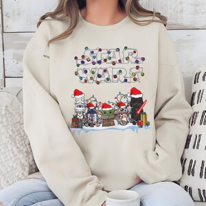 Pode incluir: Camisola bege com a inscri&ccedil;&atilde;o "STAR WARS" num design festivo com luzes coloridas. Abaixo, personagens ic&oacute;nicos como Stormtrooper, R2-D2, Grogu e Darth Vader s&atilde;o retratados com gorros de Pai Natal, rodeados de presentes.