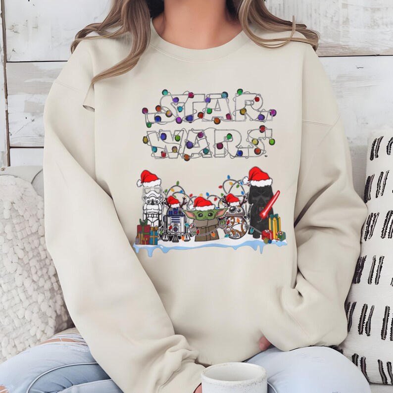 Pode incluir: Camisola bege com a inscri&ccedil;&atilde;o "STAR WARS" num design festivo com luzes coloridas. Abaixo, personagens ic&oacute;nicos como Stormtrooper, R2-D2, Grogu e Darth Vader s&atilde;o retratados com gorros de Pai Natal, rodeados de presentes.