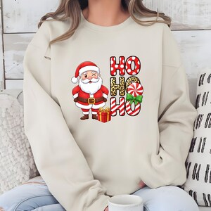 Op de afbeelding: Beige sweatshirt met een feestelijk ontwerp. Het heeft een knipogende kerstman, een rode geschenkdoos en de woorden "HO HO HO" in rode stippen, luipaardprint en zuurstokstrepen, met een groene strik.