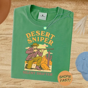 以下が含まれることがあります： 「Desert Sniper」の文字と、ライフルを構えるカウボーイのグラフィックがプリントされたグリーンのTシャツ。「Silent Drifter」と「Ships Fast！」の文字もプリントされています。青いキャップとネックレスも確認できます。