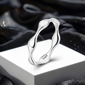 May include: A silver ring with a fluid, organic design. The ring has a smooth, polished finish and features the marking "S925" on the inside. The ring is displayed on a white surface against a dark, textured background.