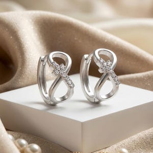 Può includere: Un paio di orecchini a cerchio in argento con un design a simbolo di infinito integrato con una piccola croce. Ogni croce è impreziosita da minuscole pietre chiare e scintillanti incastonate sulla superficie, per una finitura metallica lucida.