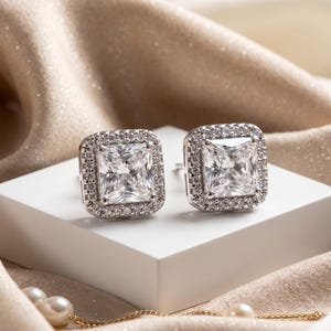 May include: A pair of silver-toned stud earrings featuring a large, square-cut clear centre stone surrounded by a halo of smaller, round-cut clear stones. The earrings are set in a metallic frame and display a brilliant, reflective finish.
