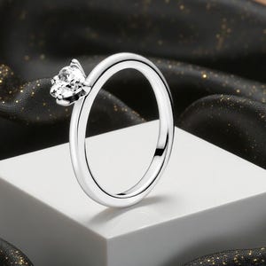 Puede incluir: Un anillo plateado con una gema transparente y brillante. El anillo se exhibe sobre una superficie blanca, con un fondo de tela negra con motas doradas. La gema es el punto focal, captando la luz.