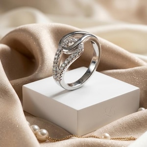 Könnte beinhalten: Ein silberfarbener Ring mit einem Bypass-Design und geteilter Ringschiene. Eine Seite des Rings ist mit kleinen, klaren, funkelnden Steinen besetzt, während die andere Seite eine glatte, polierte Metalloberfläche aufweist, was einen Schleifeneffekt erzeugt.
