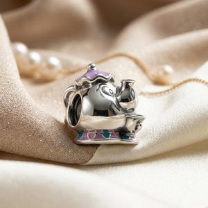 Könnte beinhalten: Ein silberner Charm in Form einer Teekanne mit einem lächelnden Gesicht, einem hellvioletten Deckel und einer Basis mit abwechselnden hellvioletten und türkisfarbenen Emaille-Akzenten. Der Charm hat eine polierte Metalloberfläche und eine hohle Mitte.
