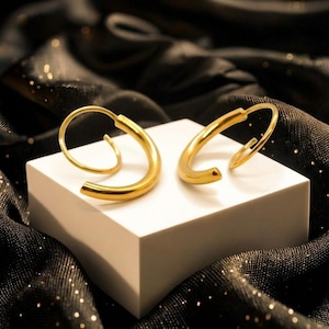 May include: A pair of gold-colored hoop earrings with a curved design. The earrings are displayed on a white cube against a backdrop of black fabric.