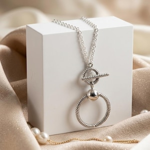 Puede incluir: Un collar de tono plateado con una cadena fina y un cierre de barra. El colgante consta de un anillo circular texturizado conectado a una esfera plateada lisa y pulida, la cual está unida a un mecanismo de cierre de barra horizontal.