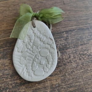 Op de afbeelding: Een lichtbeige, eivormig ornament met een getextureerd, patroonachtig oppervlak. Het is versierd met een groen lint en touw om op te hangen. Het ornament is waarschijnlijk handgemaakt en geschikt voor woondecoratie of seizoensgebonden displays.