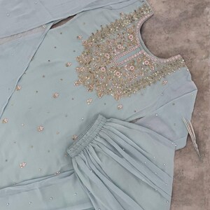 Op de afbeelding: Lichtblauw geborduurd salwar kameez-ensemble. De top heeft ingewikkelde gouden en roze borduursels rond de halslijn en verspreide bloemmotieven. De set bevat een bijpassende broek en een doorschijnende dupatta, allemaal versierd met kleine, sprankelende details.