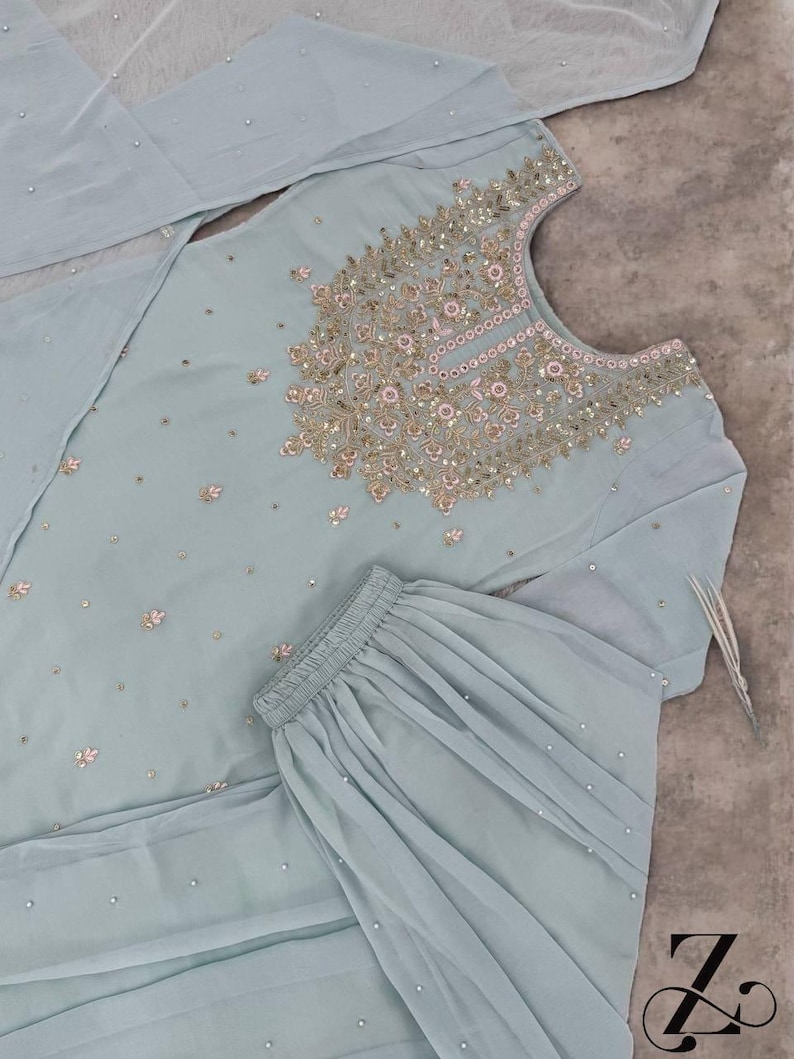 Op de afbeelding: Lichtblauw geborduurd salwar kameez-ensemble. De top heeft ingewikkelde gouden en roze borduursels rond de halslijn en verspreide bloemmotieven. De set bevat een bijpassende broek en een doorschijnende dupatta, allemaal versierd met kleine, sprankelende details.