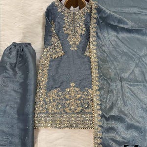 May include: A three-piece, gray-blue embroidered outfit. The set includes a long-sleeved tunic, matching pants, and a sheer scarf. The tunic and scarf are embellished with gold embroidery and small, scattered embellishments. The outfit is displayed on a wooden hanger.