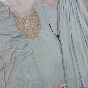 Op de afbeelding: Een lichtblauwe, driedelige geborduurde outfit. De tuniek met lange mouwen en de bijpassende rok zijn versierd met ingewikkelde gouden en zilveren borduursels en kleine, verspreide versieringen. Een doorschijnende sjaal maakt de set compleet.