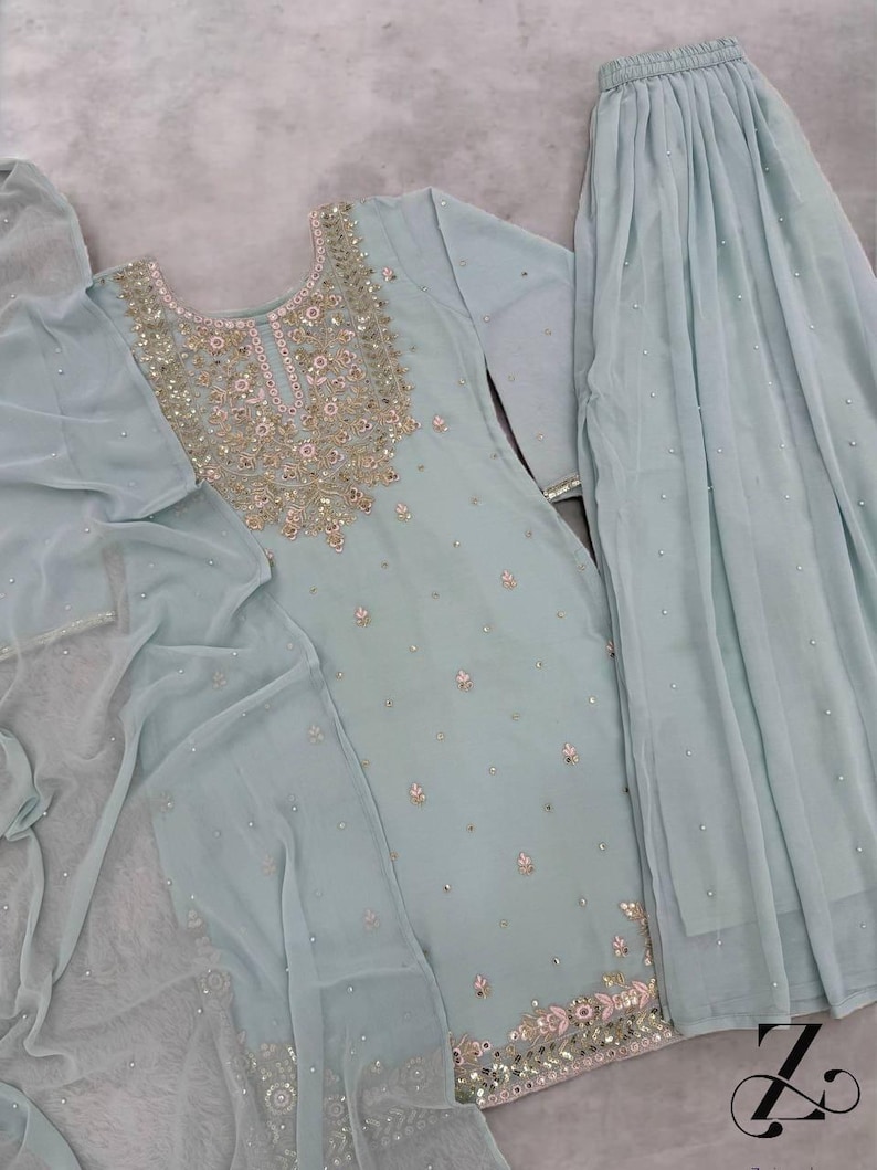 Op de afbeelding: Een lichtblauwe, driedelige geborduurde outfit. De tuniek met lange mouwen en de bijpassende rok zijn versierd met ingewikkelde gouden en zilveren borduursels en kleine, verspreide versieringen. Een doorschijnende sjaal maakt de set compleet.