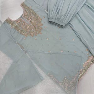 Op de afbeelding: Een lichtblauwe salwar kameez set met gedetailleerde borduursels. De top is versierd met gouden en roze borduursels rond de halslijn en zoom, met verspreide versieringen. De set bevat een bijpassende broek en een doorzichtige dupatta. De outfit ligt op een witte ondergrond.