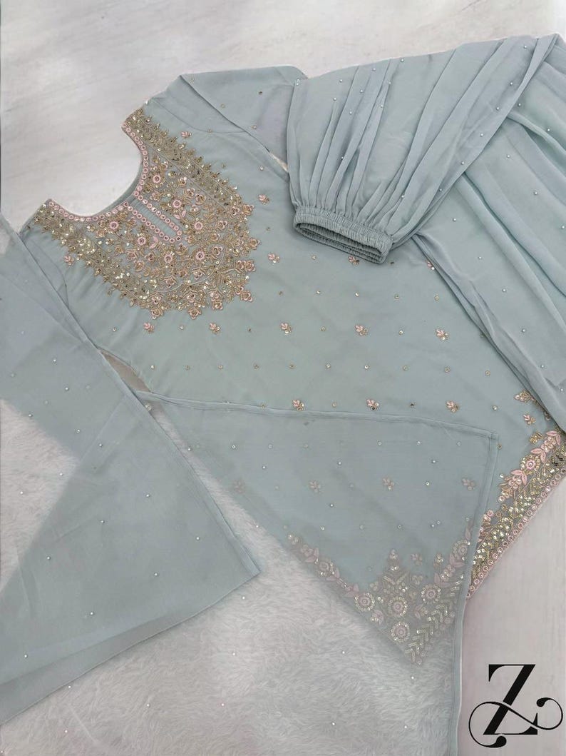 Op de afbeelding: Een lichtblauwe salwar kameez set met gedetailleerde borduursels. De top is versierd met gouden en roze borduursels rond de halslijn en zoom, met verspreide versieringen. De set bevat een bijpassende broek en een doorzichtige dupatta. De outfit ligt op een witte ondergrond.