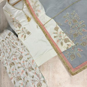 Puede incluir: Conjunto salwar kameez bordado de color marfil. El conjunto incluye una túnica, un pantalón y un dupatta transparente. La túnica y el pantalón presentan bordados florales en colores pastel. El dupatta tiene un borde rosa con ribete dorado.