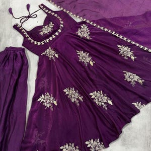 May include: A deep purple formal dress set with matching trousers and a scarf. The dress features floral embroidery with gold accents. The dress has a scoop neckline and spaghetti straps. The set is made of a shiny, flowing fabric.
