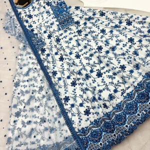 May include: White and blue floral embroidered dress with matching sheer scarf and white pants. The dress features intricate blue floral patterns and detailed embroidery along the edges. The scarf has a delicate floral design.