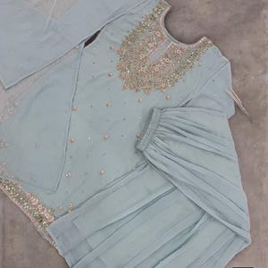 Op de afbeelding: Een lichtblauwe, versierde formele outfit. De set bevat een tuniek met lange mouwen en ingewikkelde borduursels rond de halslijn en verspreide versieringen. De bijpassende broek heeft een gelaagd ontwerp. Een doorschijnende sjaal maakt de set compleet.