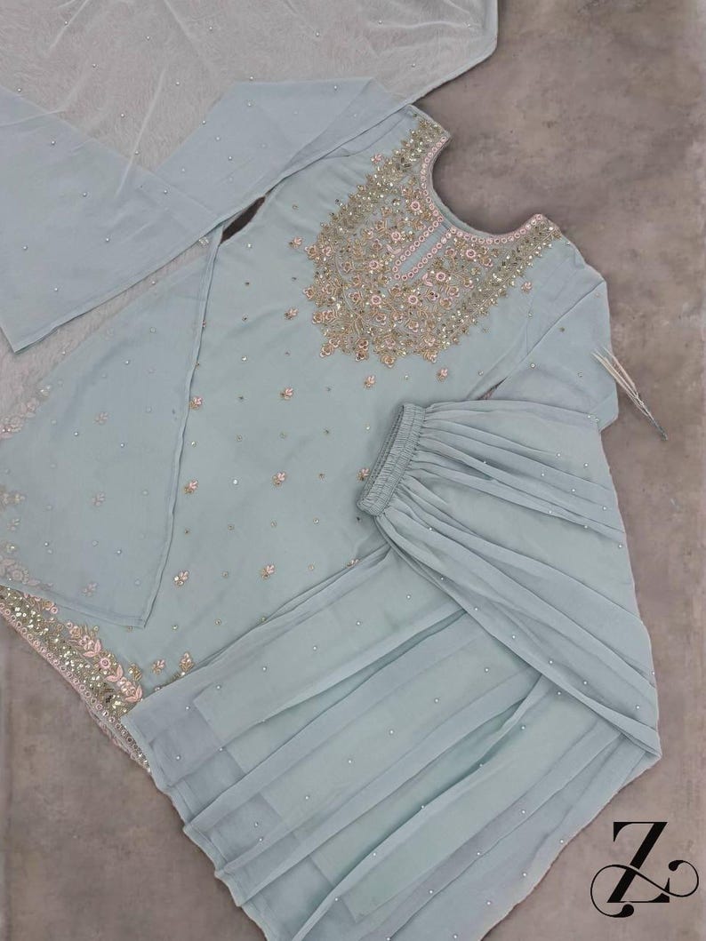 Op de afbeelding: Een lichtblauwe, versierde formele outfit. De set bevat een tuniek met lange mouwen en ingewikkelde borduursels rond de halslijn en verspreide versieringen. De bijpassende broek heeft een gelaagd ontwerp. Een doorschijnende sjaal maakt de set compleet.