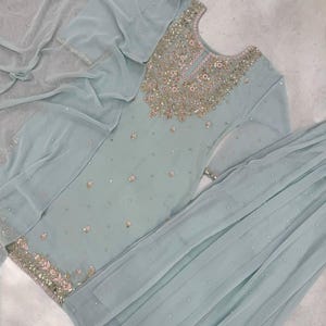 Op de afbeelding: Een lichtblauwe, versierde salwar kameez set. De top heeft ingewikkelde borduursels rond de halslijn en zoom, met verspreide versieringen. De set bevat een bijpassende dupatta en broek, waardoor een samenhangend ensemble ontstaat. De stof lijkt licht en vloeiend.