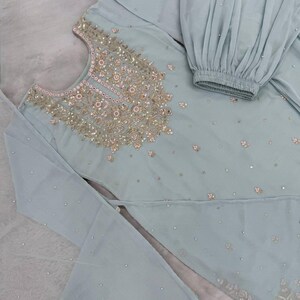 Op de afbeelding: Een lichtblauwe, versierde salwar kameez set. De top heeft ingewikkelde gouden en roze borduursels rond de halslijn en verspreide versieringen. De set bevat een top met uitlopende mouwen en een bijpassende broek met een geplooide manchet.
