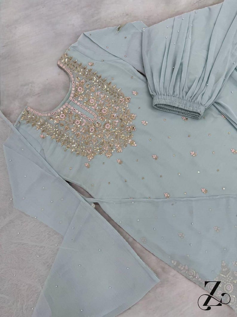 Op de afbeelding: Een lichtblauwe, versierde salwar kameez set. De top heeft ingewikkelde gouden en roze borduursels rond de halslijn en verspreide versieringen. De set bevat een top met uitlopende mouwen en een bijpassende broek met een geplooide manchet.
