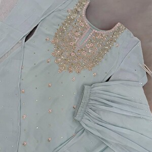 Op de afbeelding: Een lichtblauwe, tweedelige geborduurde outfit. De top heeft ingewikkelde gouden en roze versieringen rond de halslijn en verspreid over de stof. De bijpassende broek heeft een uitlopend ontwerp. De outfit is versierd met kleine, sprankelende accenten.