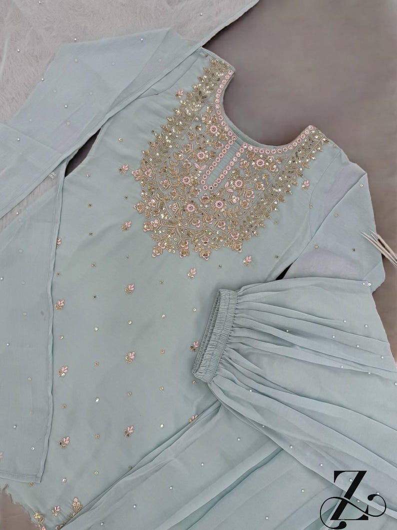 Op de afbeelding: Een lichtblauwe, tweedelige geborduurde outfit. De top heeft ingewikkelde gouden en roze versieringen rond de halslijn en verspreid over de stof. De bijpassende broek heeft een uitlopend ontwerp. De outfit is versierd met kleine, sprankelende accenten.