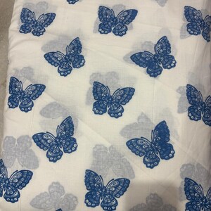 May include: A white fabric featuring a repeating pattern of blue butterflies. The design includes both vibrant, fully colored blue butterflies and faint, light blue shadow-like butterfly silhouettes printed across the textile surface.