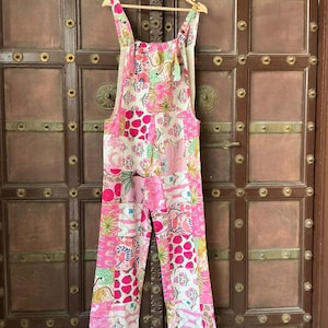 Op de afbeelding: Een roze en witte jumpsuit met bloemenprint en verstelbare bandjes hangt aan een houten hanger. De jumpsuit heeft een patchwork-ontwerp met verschillende bloempatronen en een silhouet met wijde pijpen. De achtergrond is een donkere houten deur.