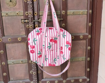 Puede incluir: Un bolso tote a rayas rosas y blancas con un estampado de cerezas. El bolso tiene forma rectangular con un pequeño bolsillo en la parte delantera y asas largas. Cuelga de una puerta de madera marrón.