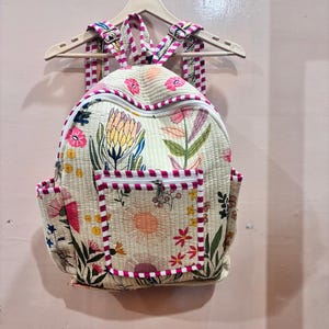 May include: A quilted backpack in a cream colour featuring a vibrant floral print with pink, yellow, and green botanical motifs. The bag includes a front zipped pocket, side pockets, and adjustable shoulder straps with a pink and white striped trim.
