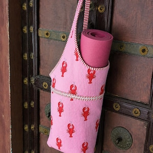 May include: A pink yoga mat bag featuring a repeating red lobster pattern and white and red striped trim. The bag has a long, matching striped shoulder strap and contains a rolled-up solid dark pink yoga mat inside the cylindrical fabric carrier.