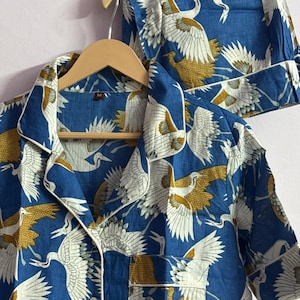 May include: A blue pajama set featuring a repeating pattern of white and gold cranes in flight. The short-sleeved button-down shirt has a notched collar with white piping, a single chest pocket, and a size M tag. Matching shorts are included.