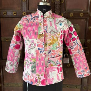 May include: A quilted jacket with a stand-up collar, featuring a patchwork design in pink, white, and floral patterns. The jacket has long sleeves and two front pockets. The design includes images of flowers, strawberries, and a leopard.