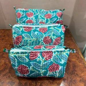 May include: Three rectangular quilted pouches featuring a teal background with a repeating pattern of pink flowers and light blue leaves. Each pouch has a white zipper closure along the top edge and a small fabric tab on the side for easy handling.