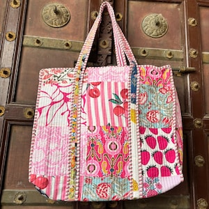Può includere: Una borsa tote patchwork con una varietà di motivi rosa e bianchi, tra cui strisce, disegni floreali e motivi di frutta. La borsa ha una texture trapuntata e manici robusti, perfetta per trasportare oggetti.