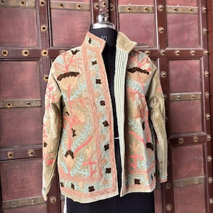 Puede incluir: Chaqueta beige con bordados florales en tonos melocotón, marrón y turquesa. La chaqueta tiene un cuello alto y se exhibe en un maniquí negro. El forro interior es a rayas blancas y verdes. Prenda de vestir única.
