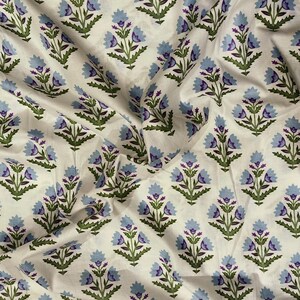 Puede incluir: Tela blanca con un estampado floral repetitivo. El diseño presenta flores azules estilizadas con centros morados y hojas verdes. La tela está arrugada, lo que sugiere que es un textil.