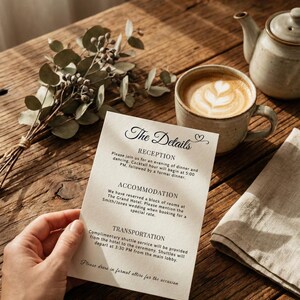 May include: A wedding invitation with the words "The Details" in elegant script. The invitation is held by a hand and rests on a wooden table, alongside a cup of coffee with latte art, a teapot, and a bouquet of greenery.