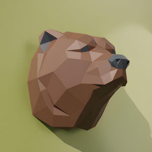Könnte beinhalten: Eine braune, geometrische Bärenkopfskulptur mit polygonalem Design. Die Nase des Bären ist dunkelgrau, und die Skulptur wirft einen Schatten auf den hellgrünen Hintergrund. Der Bärenkopf ist leicht nach rechts geneigt.