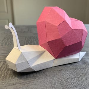 Peut inclure: Une sculpture d'escargot en papier géométrique. La coquille est en forme de cœur rose à facettes. Le corps est blanc avec des facettes géométriques. Les antennes sont blanches et incurvées.