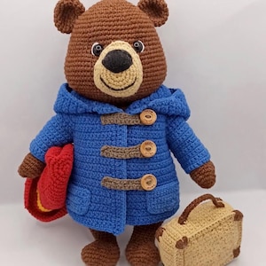 Puede incluir: Un oso de ganchillo hecho a mano con un abrigo azul con cierres de madera, un sombrero rojo y una maleta beige. El oso tiene una cabeza marrón, un hocico beige y ojos negros. El abrigo tiene capucha y bolsillos.