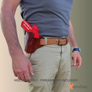 May include: A brown leather holster with a red training gun, worn on a light brown belt. The text "FIREARM NOT INCLUDED" is visible on the red gun. The text "HOLSTER ONLY - FIREARM NOT INCLUDED" is at the bottom.